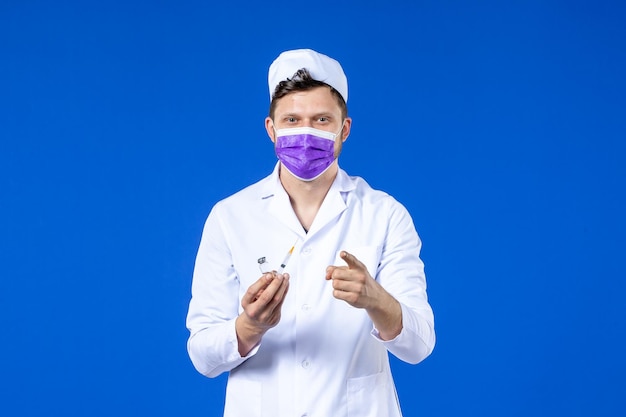 Front view of male doctor in medical suit and mask with injection and vaccine on blue 