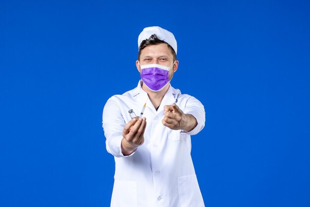 Front view of male doctor in medical suit and mask with injection and vaccine on blue 