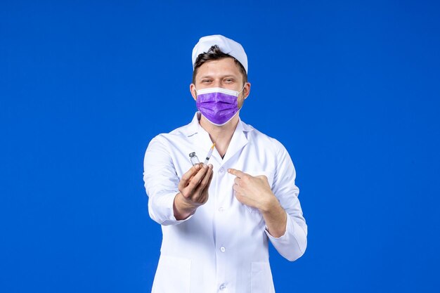 Вид спереди мужского врача в медицинском костюме и маске с инъекцией и вакциной на синем
