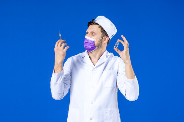 Front view of male doctor in medical suit and mask with injection and vaccine on blue 