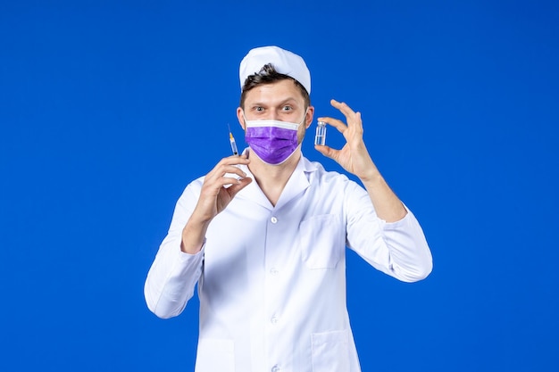 Front view of male doctor in medical suit and mask with injection and vaccine on blue 