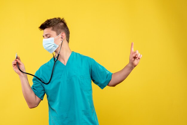 Front view of male doctor in medical suit and mask with stethoscope on the yellow wall