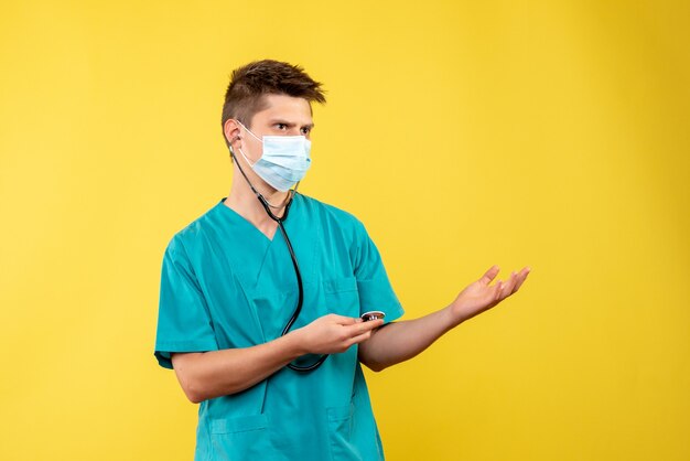 Front view of male doctor in medical suit and mask with stethoscope on yellow wall