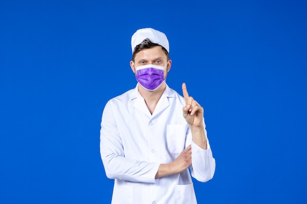 Front view of male doctor in medical suit and purple mask counting on blue 