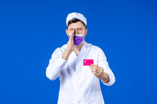 Front view of male doctor in medical suit and purple mask holding credit card on blue 