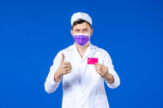 Front view of male doctor in medical suit and purple mask holding credit card on blue 