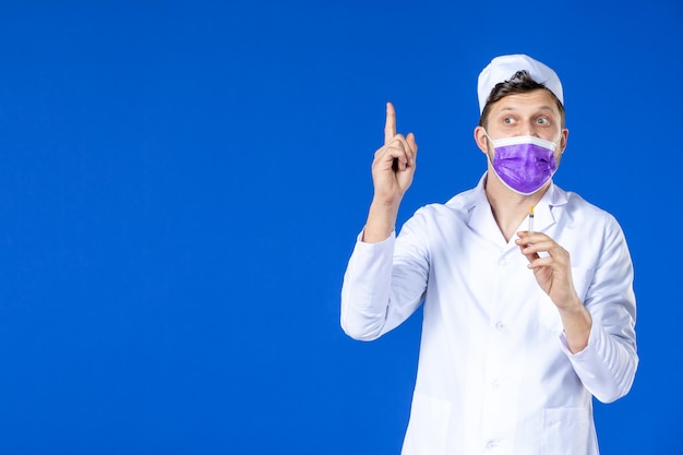 Front view of male doctor in medical suit and purple mask holding injection on blue 