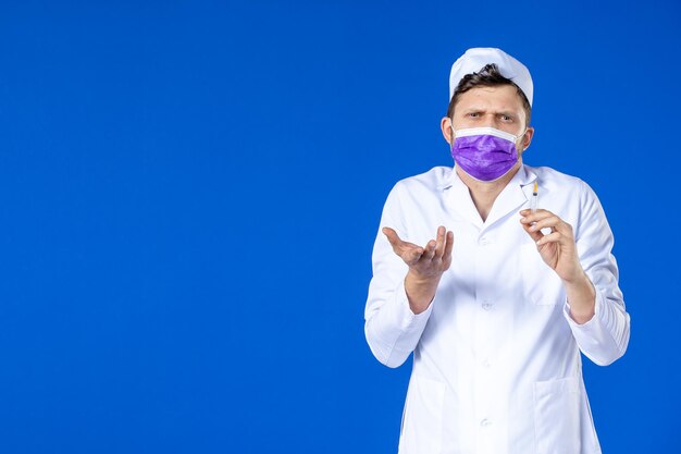 Front view of male doctor in medical suit and purple mask holding injection on blue 