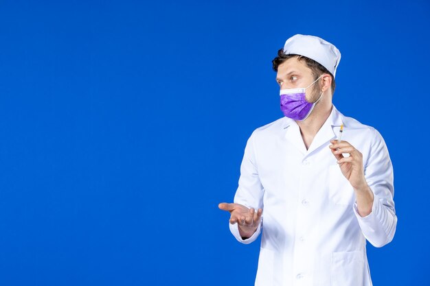 Front view of male doctor in medical suit and purple mask holding injection on blue 