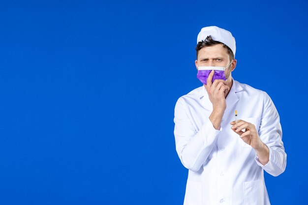 Front view of male doctor in medical suit and purple mask holding injection on blue 