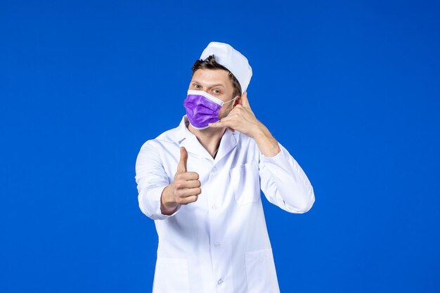 Вид спереди мужчины-врача в медицинском костюме и фиолетовой маске, имитирующей телефонный звонок на синем