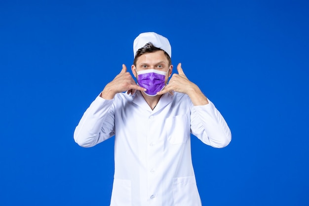 医療スーツと紫色のマスクで電話ポーズオンブルーを示す男性医師の正面図