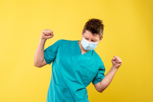 Вид спереди мужчины-врача в медицинском костюме и стерильной маске, радующегося на желтой стене
