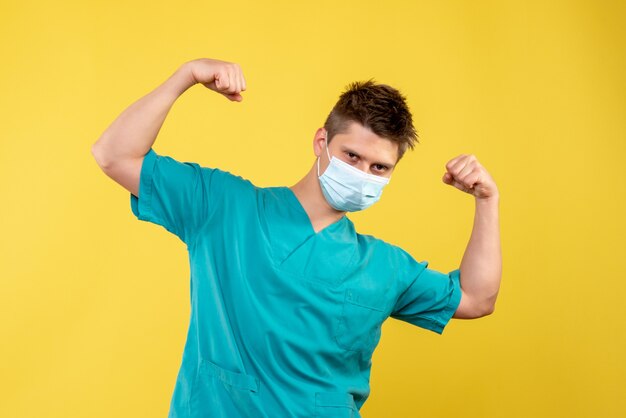 Вид спереди мужского врача в медицинском костюме и стерильной маске на желтой стене
