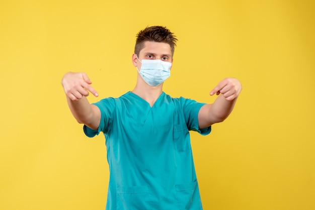Вид спереди мужского врача в медицинском костюме и стерильной маске на желтой стене