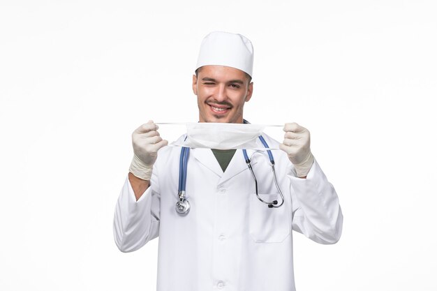 Front view male doctor in medical suit and wearing a mask against coronavirus on the white desk virus covid- illness disease pandemic