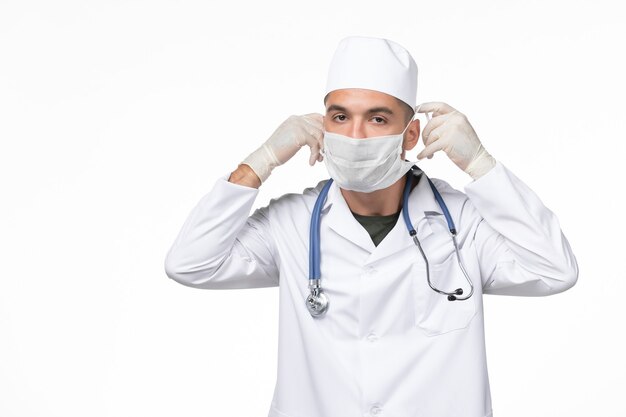 Front view male doctor in medical suit and wearing a mask against coronavirus on white wall virus covid- illness disease pandemic