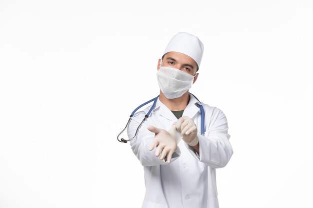 Front view male doctor in medical suit and wearing a mask against covid on light-white wall covid virus medicine pandemic