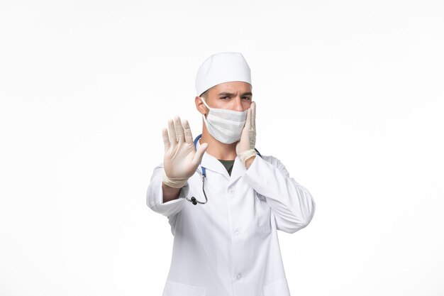Front view male doctor in medical suit and wearing a mask against covid on the light-white wall covid virus medicine pandemic