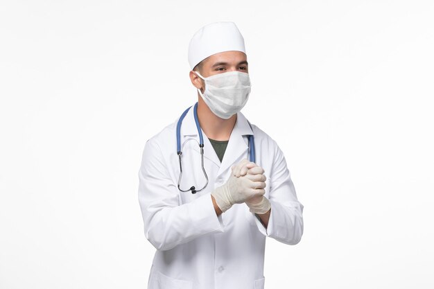 Front view male doctor in medical suit and wearing a mask against covid on light white wall virus covid- illness disease pandemic