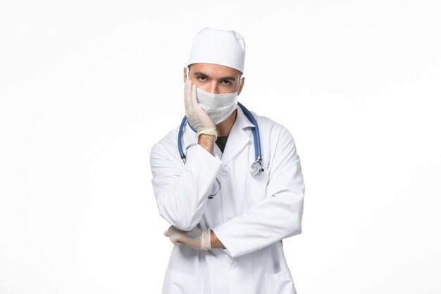 Front view male doctor in medical suit and wearing a mask against covid thinking on white wall covid illness disease pandemic virus