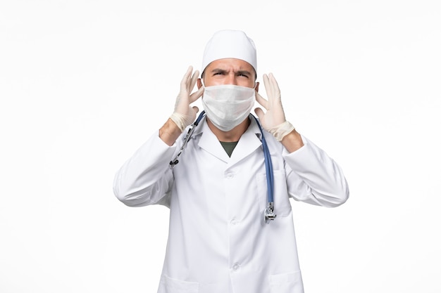 医療スーツを着て、白い机の上に青い聴診器でcovidに対してマスクを身に着けている正面図男性医師covid病気パンデミックウイルス