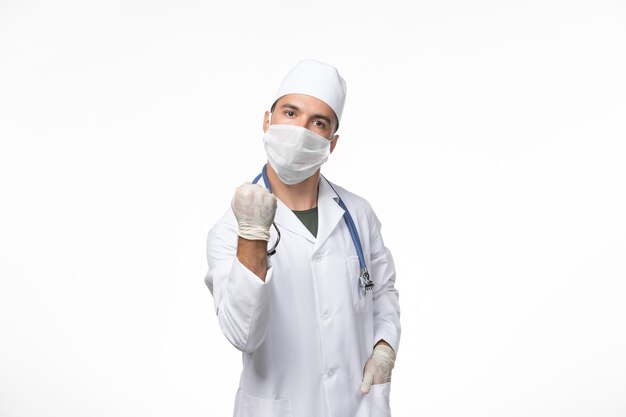 Front view male doctor in medical suit and wearing a mask against covid with blue stethoscope on white wall covid- illness disease pandemic virus