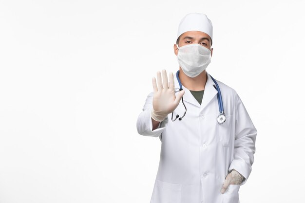 Front view male doctor in medical suit and wearing a mask against covid with stethoscope on light-white desk virus covid- illness disease pandemic