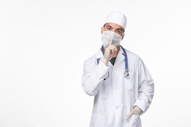 Front view male doctor in medical suit and wearing a mask against covid with stethoscope on light white wall virus covid- illness disease pandemic