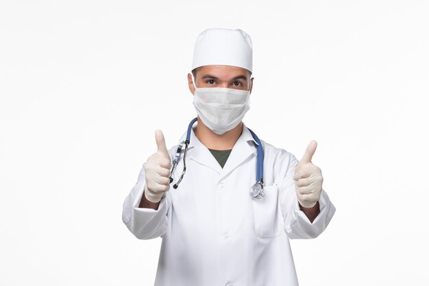 Front view male doctor in medical suit and wearing a mask against covid with stethoscope posing on white wall virus covid- illness disease pandemic