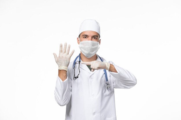 Front view male doctor in medical suit and wearing a mask against covid with stethoscope on the white desk covid- illness disease pandemic virus