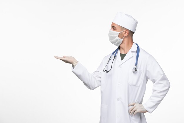 Front view male doctor in medical suit and wearing a mask against covid with stethoscope on the white wall virus covid- illness disease pandemic