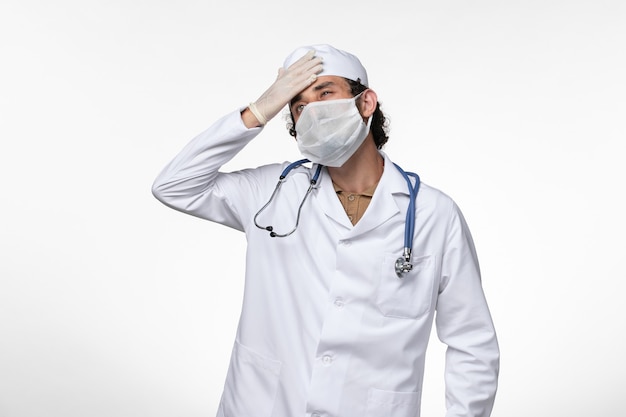 医療スーツを着て、白い壁の病気のウイルス感染症に頭痛を持っていることからの保護としてマスクを身に着けている正面図の男性医師