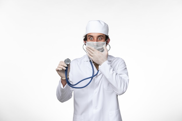 Vista frontale medico maschio in tuta medica e indossa una maschera come protezione da covid- utilizzando uno stetoscopio sulla scrivania bianca virus malattia covid- pandemia