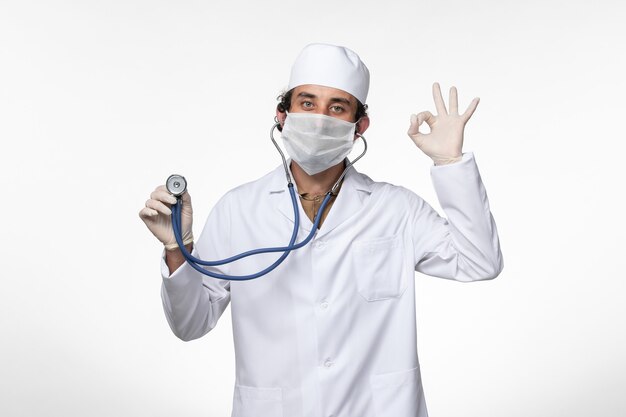 Front view male doctor in medical suit and wearing a mask as a protection from covid- using a stethoscope on the white wall illness covid- pandemic