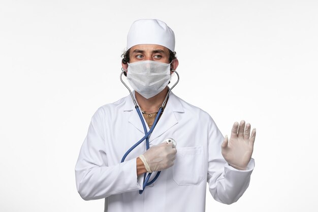 医療スーツを着て、白い壁の病気のウイルス感染症に聴診器を使用することからの保護としてマスクを着用している正面図の男性医師