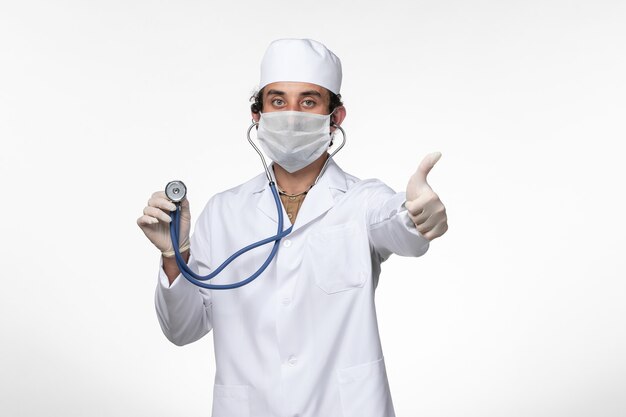 Front view male doctor in medical suit and wearing a mask as a protection from covid- using a stethoscope on white wall virus illness covid- pandemic