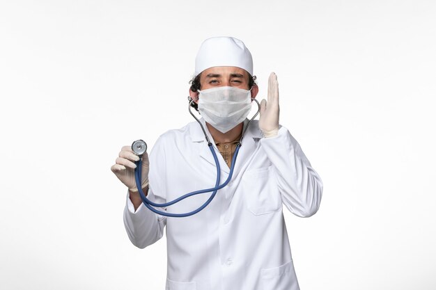 Front view male doctor in medical suit and wearing a mask as a protection from covid- using a stethoscope on white wall virus illness covid pandemic