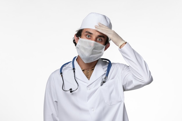 Front view male doctor in medical suit and wearing a mask as a protection from covid- on the white wall illness virus covid- pandemic