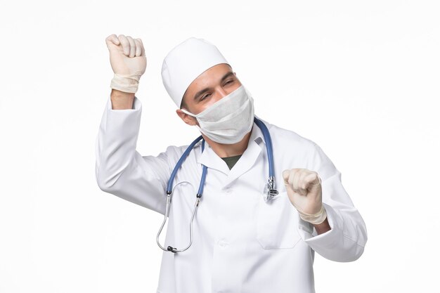 Front view male doctor in medical suit and wearing mask due to covid- dancing on white wall covid- virus pandemic virus disease