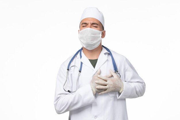 医療スーツを着て、白い壁に心臓の痛みがあるためにマスクを着用している正面図男性医師covid-ウイルスパンデミックウイルス病