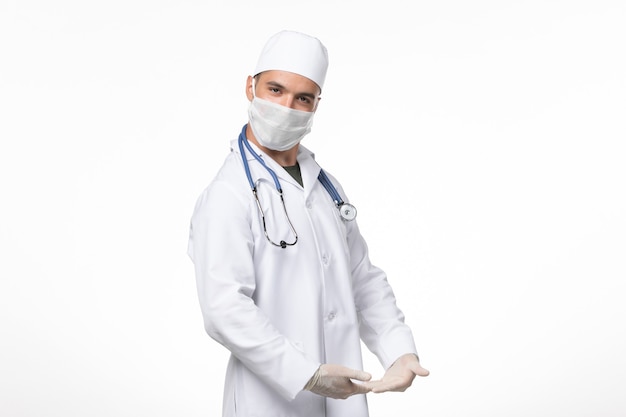 Front view male doctor in medical suit and wearing mask due to covid- posing on white wall covid- virus pandemic virus disease