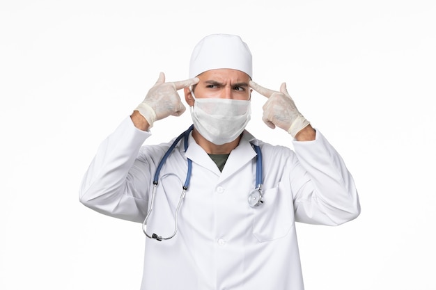 Front view male doctor in medical suit and wearing mask due to covid- thinking on white wall covid- pandemic virus disease