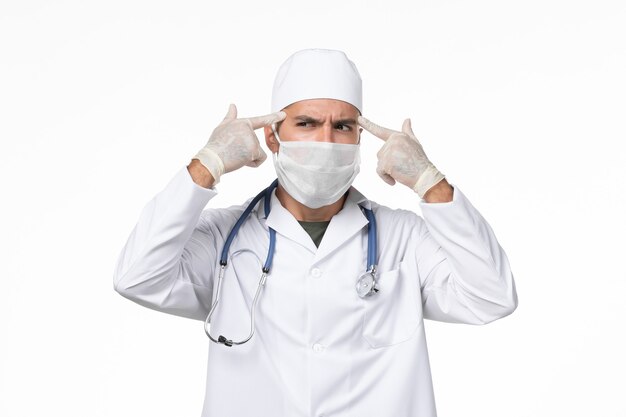 Front view male doctor in medical suit and wearing mask due to covid- thinking on white wall covid- pandemic virus disease