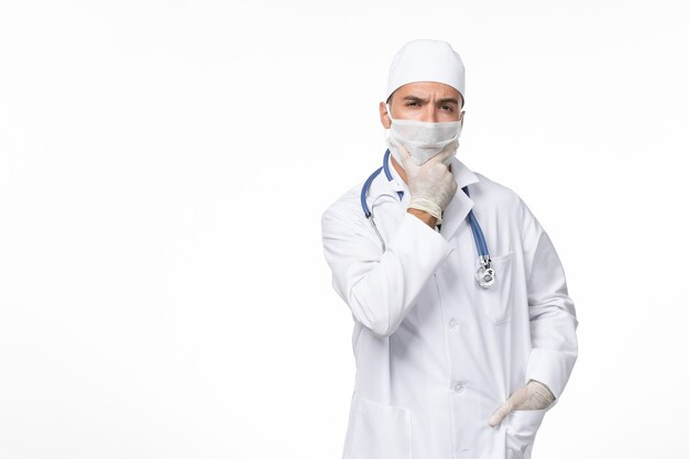 Front view male doctor in medical suit and wearing mask due to covid- thinking on white wall illness virus covid- pandemic disease