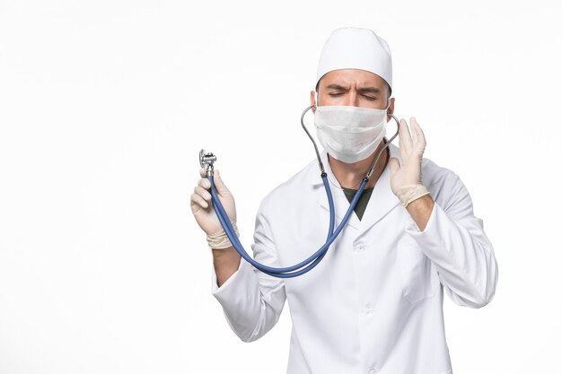 Front view male doctor in medical suit and wearing mask due to covid- using a stethoscope on white desk covid- pandemic virus disease