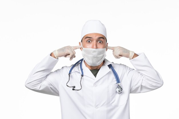 Front view male doctor in medical suit and wearing mask due to covid- on the white wall illness covid- pandemic disease