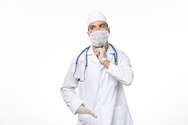 Front view male doctor in medical suit and wearing mask due to covid- on white wall illness virus covid- pandemic disease