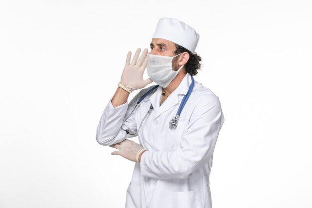 白い壁のスプラッシュウイルスコロナウイルスパンデミックを呼びかけるcovidからの保護として滅菌マスクを身に着けている医療スーツの正面図男性医師