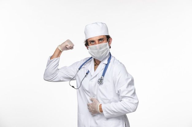 Front view male doctor in medical suit wearing sterile mask as a protection from covid flexing on white wall splash virus coronavirus pandemic health